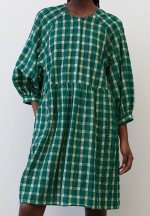 Vrouw draagt een knie-lange jurk met groene en witte ruiten, lange pofmouwen en een gerimpelde taille, staand tegen een effen achtergrond.