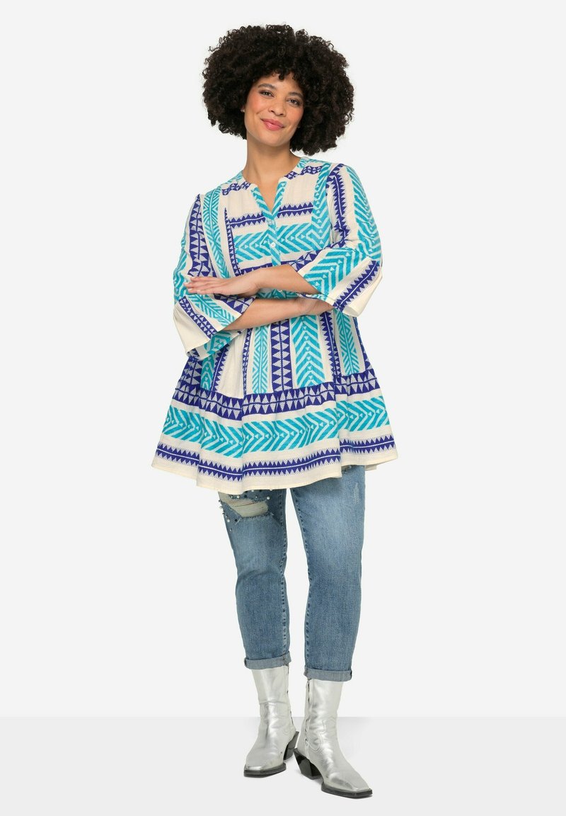 Kolorowa tunika z geometrycznymi niebieskimi i białymi wzorami, w połączeniu z jasnymi dżinsami. Wyjątkowe srebrne botki za kostkę o eleganckim designie.