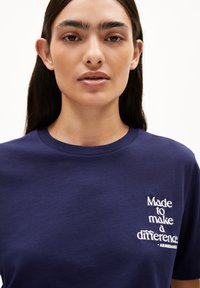 Frau mit langen dunklen Haaren, die ein navyblaues T-Shirt mit dem weißen Text "Made to make a difference - ARMEDANGELS" auf der Brust trägt.