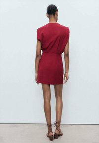 Short red dress with cap sleeves, cinched waist, and a straight cut. Smooth fabric and above-the-knee length. Brown strappy heeled sandals.
