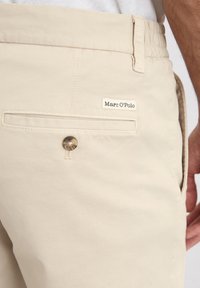 Beige shorts made of cotton blend fabric, featuring a back pocket with a button and a small brand label with "Marc O'Polo" on it.