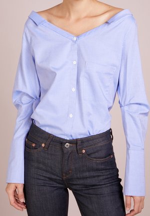 Femme portant une chemise bleu clair à épaules dénudées avec boutons, rentrée dans un jean denim bleu foncé, avec manches longues et poche poitrine.