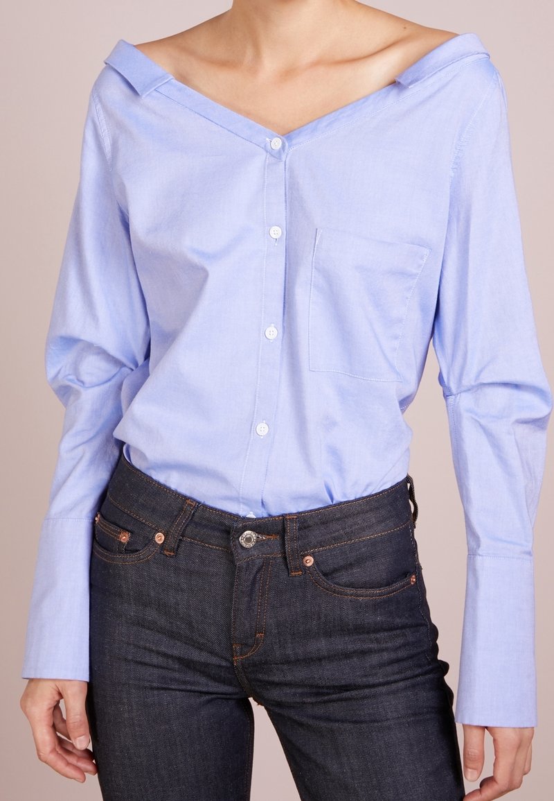Femme portant une chemise bleu clair à épaules dénudées avec boutons, rentrée dans un jean denim bleu foncé, avec manches longues et poche poitrine.