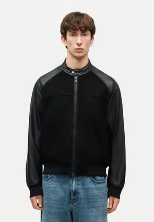 Νεαρός άνδρας που φοράει μαύρο μπουφάν με φερμουάρ και δερμάτινα μανίκια, με γιακά με κουμπιά, συνδυασμένο με μπλε τζιν παντελόνι, στέκεται μπροστά σε απλό φόντο.