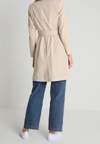 Trench de color beige con cintura ceñida, mangas largas y un diseño aerodinámico. Combinado con jeans de pierna ancha azules y zapatillas blancas.