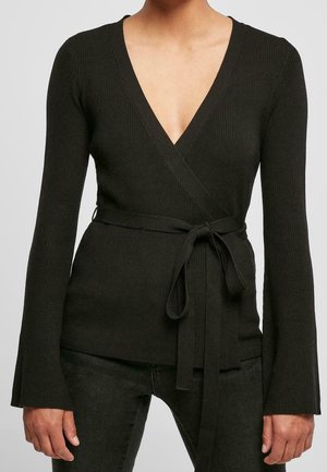 Femme portant un haut cache-cœur en maille côtelée noire avec manches longues et ceinture nouée à la taille, associé à un pantalon noir.