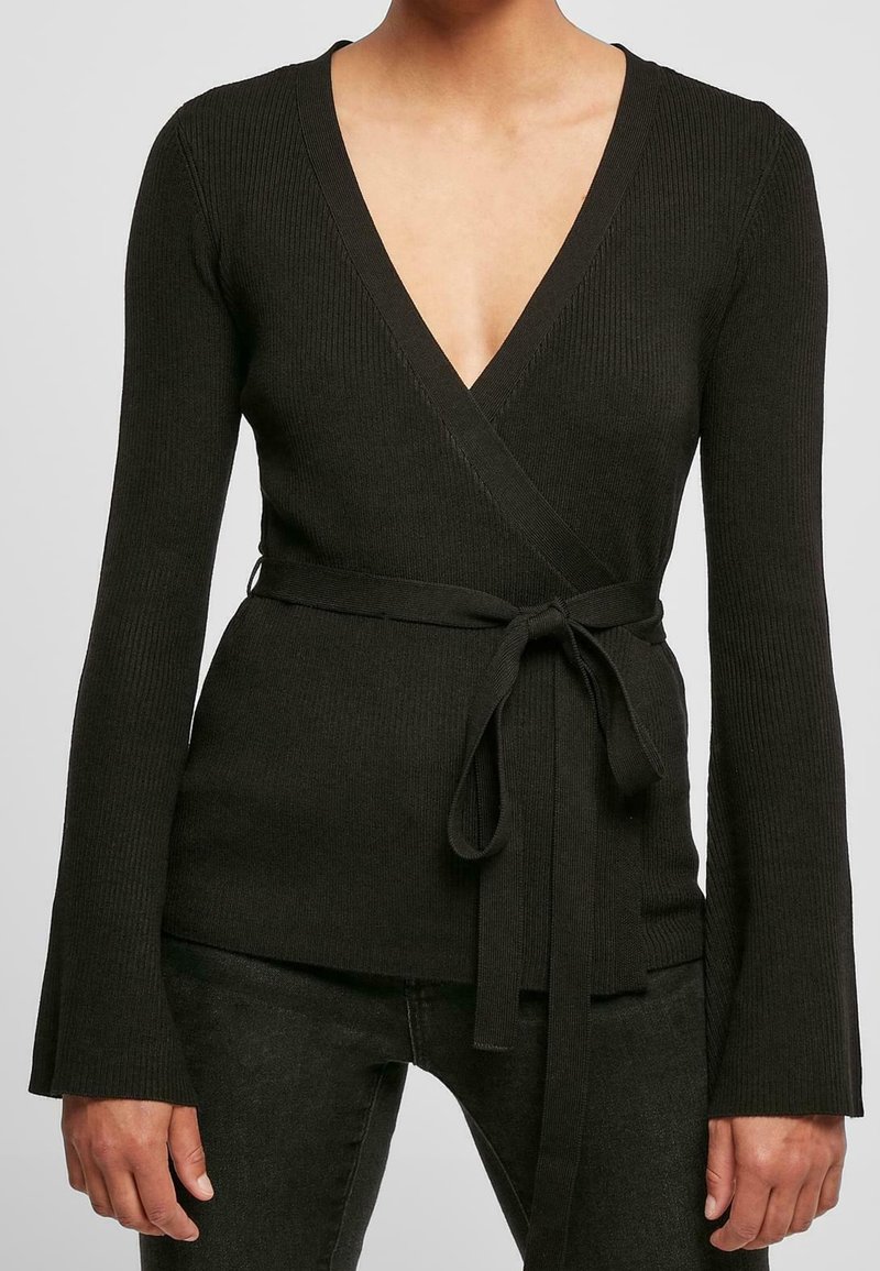 Femme portant un haut cache-cœur en maille côtelée noire avec manches longues et ceinture nouée à la taille, associé à un pantalon noir.