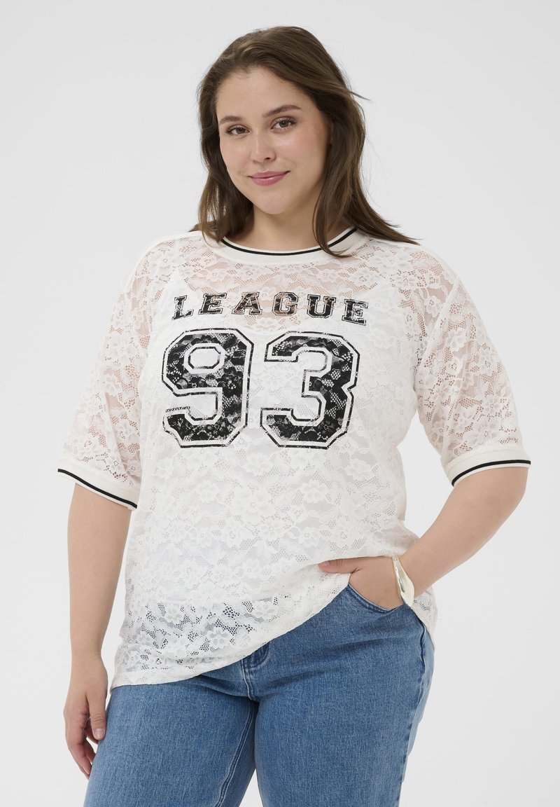 Femme portant une chemise en dentelle blanche avec le texte « LEAGUE 93 » et un jean bleu, une main dans la poche, souriant devant un fond uni.