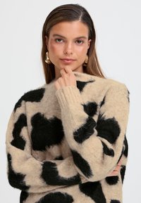 Pull beige avec un grand imprimé vache noir, fabriqué en matériau doux et texturé. Poignets et ourlet côtelés, manches longues et col rond.
