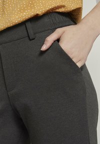 Donkergrijze broek van een gestructureerde stof, met een zijzak en een elastische tailleband. De hand van het model zit in de zak.
