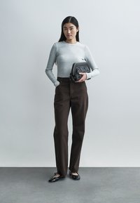 Graues geripptes Langarmoberteil, braune hoch taillierte Hose, schwarze Handtasche mit Nietenakzenten und schwarze Ballerinas. Modell steht vor einem hellen Hintergrund.