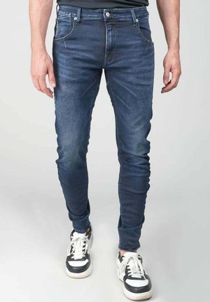 Jeans oscuros de corte ajustado con diseño de cinco bolsillos, fabricados en denim. Combinados con zapatillas negras y blancas. Textura suave, con un desvanecimiento mínimo.