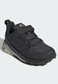 Zapatilla deportiva negra con parte superior de malla, superposiciones sintéticas, correa ajustable y suela de goma con dibujo texturizado para un mejor agarre.