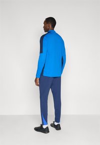 Modrý atletický dlhý rukáv s námorníckymi detailmi, skombinovaný s námorníckymi zúženými nohavicami so zipsami na boku a čiernymi športovými topánkami.