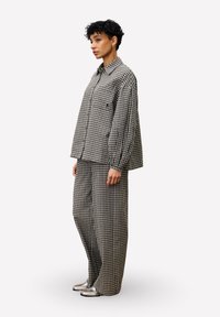 Vrouw met kort krullend haar, gekleed in een zwart-wit geruite oversize blouse en wijde broek, in profiel staand met zilveren schoenen.