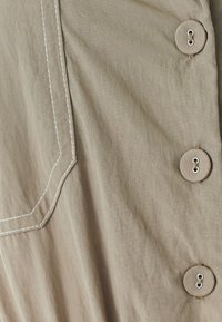 Béžový látkový oděv s výraznou kapsou a kontrastním bílým stehem, doplněný čtyřmi velkými, kulatými knoflíky na boku.