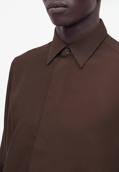 Gros plan d'une personne portant une chemise marron boutonnée avec un col pointu sur un fond uni.