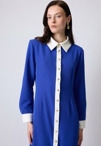 Blue long-sleeve dress featuring a white collar and button placket with gold buttons. Smooth fabric texture and fitted silhouette.