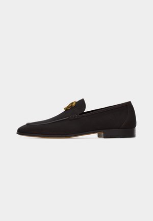 Black leather loafer with low heel and decorative gold emblem on the upper, shown from the side on a white background.