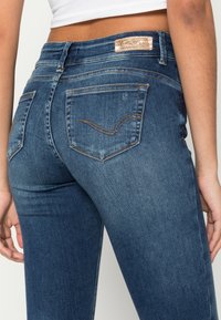 Calças de ganga azul-escuro com um corte justo, apresentando bolsos traseiros com costura curva e uma etiqueta metálica da marca na cintura.