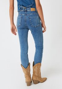 Vaqueros de denim azul ajustados con cintura de talle medio, bolsillos traseros y una etiqueta de cuero marrón. Combinados con botas de vaquero beige con costuras decorativas.