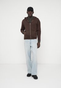 Barna cipzáras kapucnis felső egy zöld kockás inget viselve, világoskék bővített farmernadrággal és fekete cipővel párosítva. Egyszerű háttér.