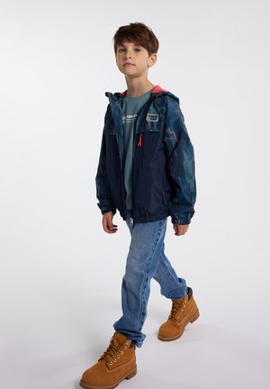 Ragazzo che cammina indossando una giacca blu mimetica, una maglietta teal Timberland, jeans blu e stivali Timberland beige su uno sfondo chiaro e semplice.