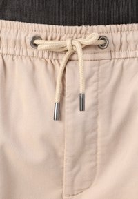 Pantalons beige à cordon de serrage avec une taille texturée gris foncé. Les caractéristiques incluent un œillet en métal et un cordon tressé couleur crème avec des embouts métalliques.