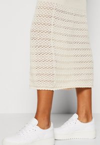 Białe trampki z teksturowanym, wytrzymałym materiałem i płaską podeszwą, zestawione z kremową dzianinową spódnicą do kolan z ażurowym wzorem.