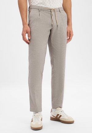 Man wearing beige slim-fit trousers with drawstring waist and white sneakers with brown stripes, standing against white background.