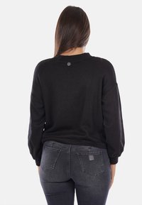 Maglione nero a maglia con maniche lunghe e larghe e scollatura rotonda. Presenta un piccolo logo sul retro. Abbinato a jeans attillati grigio scuro.