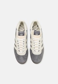 Zapatillas Adidas con un upper de ante gris, acentos en color crema, cordones blancos y una plantilla estampada con el logo de Adidas.