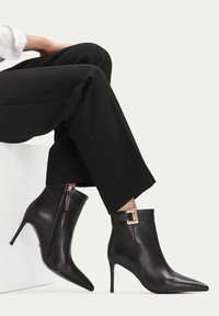 Mustad nahast terava ninaga pahkluu saapad kuldse pandlaga ja külgsipistiku kaudu, kanda mustade laia jala pükste kõrval valgel taustal.