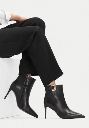 Černé kožené kotníkové boty se špičatou špičkou, zlatým sponou a bočním zipem, které jsou kombinovány s černými kalhotami s širokými nohavicemi na bílém pozadí.