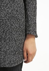 Maglione grigio a maglia testurizzata con una vestibilità ampia e polsini larghi. Primo piano del maniche e della mano, enfatizzando l'aspetto morbido del tessuto.