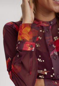 Genomskinlig bordeauxblus med blommönster i rött, orange och vitt. Har knäppning framtill och manschetter med kontrasterande knappdetaljer.