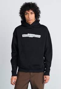 Pessoa com cabelo encaracolado, vestindo um hoodie preto com o texto "the new originals", calças castanhas, de pé contra um fundo claro e simples.