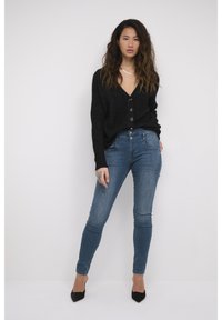 Crna pletena cardigan s dubokim V-izrezom i gumbima, uparen s plavim uskim trapericama i crnim cipelama sa špicastim vrhom. Glatke teksture svugdje.