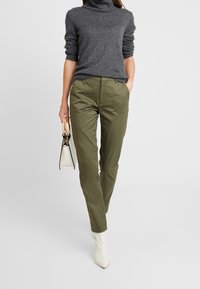 Pull en col roulé gris, pantalon vert olive, et bottes à talons blanches. Tient un petit sac à main blanc avec un accent noir.