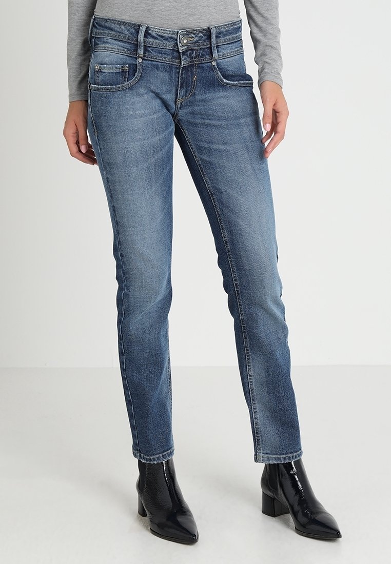 Kvinna i blå slim-fit jeans och svarta klackade ankelstövlar, stående med avslappnade händer vid sidorna.