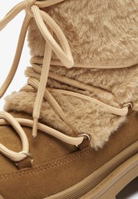Bota de imitación de piel de color marrón claro, forrada de pelaje, con una textura suave, que presenta cordones beige, ojales de metal y una suela de goma resistente para estabilidad y agarre.