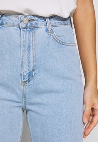 Jeans de mezclilla de tiro alto en azul claro con cremallera, botón de cobre y costuras en contraste. Cuenta con bolsillos frontales y una textura suave.