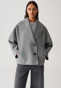 Vrouw in een oversized grijze wollen jas met brede revers, twee knopen en handen in de zakken, gecombineerd met donkergrijze jeans.