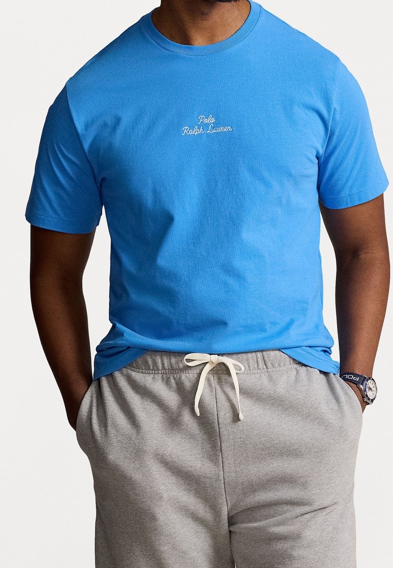 Blue cotton t-shirt with short sleeves, featuring white "Polo Ralph Lauren" embroidery at the center, paired with gray sweatpants and a watch.