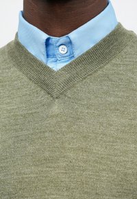 Homme portant une chemise bleu clair boutonnée sous un pull à col en V vert olive, montrant le col et la partie supérieure de la poitrine.