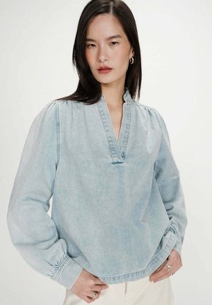 Jeune femme aux cheveux noirs et raides portant une blouse en denim bleu clair avec col mao et manches longues bouffantes, debout les mains sur les hanches.