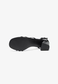 Black heeled sandal with a textured sole, featuring crossed straps on the front and a sturdy block heel. Matte finish, minimal design.