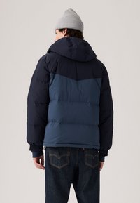 Temno modra puffarica s svetlo modrim chevron vzorom na hrbtu, z kapuco in rebrastimi manšetami. Uporabljena z temnimi jeans hlačami.