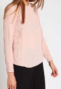 Femme portant un chemisier à manches longues rose pâle avec des détails en dentelle sur les côtés et les poignets, assorti à un pantalon noir, sur fond blanc.