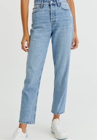 Jean taille haute bleu clair avec une jambe droite, légères distorsions et ourlet effiloché, associé à des baskets blanches. Tissu en denim texturé.